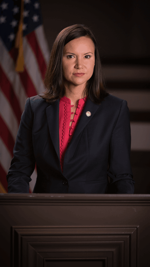 A woman in a dark suit and red blouse posed beside an American flag